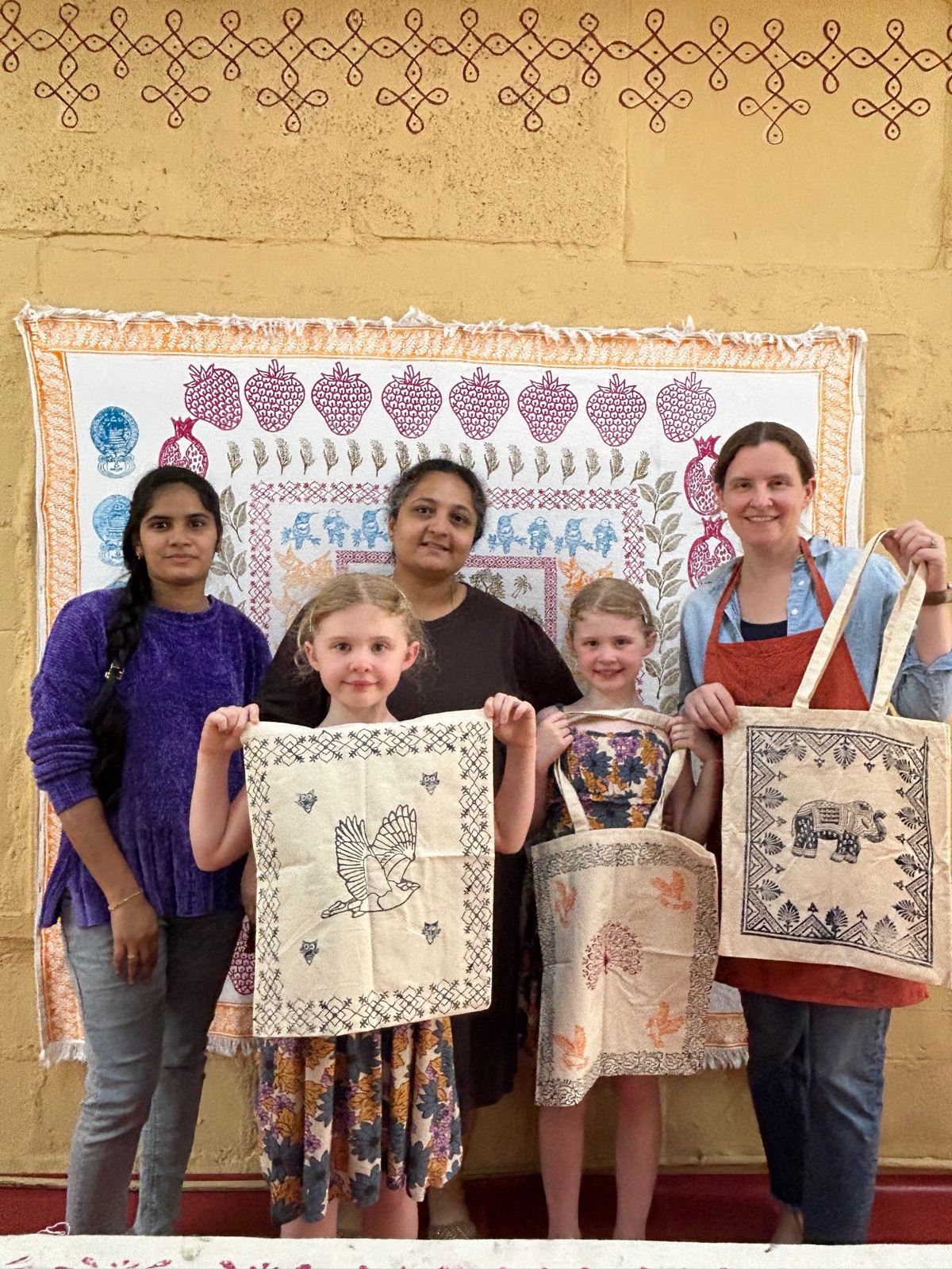 A family with two young children holding up block printed tote bags in front of a printed wall hanging